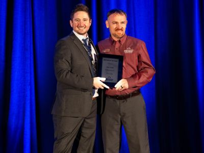 Two men standing next to each other on stage. One man presents a plaque, while the other man accepts the award